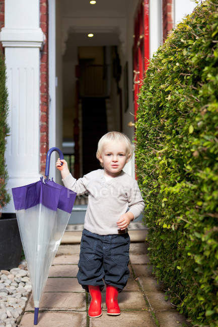 Little boy standing on front garden path with umbrella and rubber boots — Stock Photo