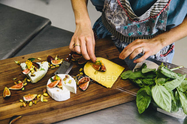 Mulher preparando prato de queijo variedade com figos, nozes, pistache, manjericão em tábua de cortar madeira — Fotografia de Stock