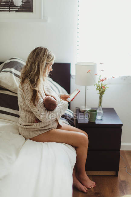 Mulher adulta sentada na cama olhando para o smartphone enquanto embala a filha recém-nascida — Fotografia de Stock