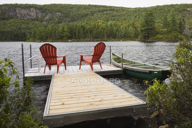 Deux chaises longues en plastique rouge Adirondack sur un quai flottant sur le lac dans la cour arrière d'une maison en bois rond de style chalet à la fin de l'été, Québec, Canada. Cette image est la propriété libérée — Photo de stock