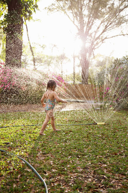 Girl in bathing costume playing in sunlit garden sprinkler water — Stock Photo