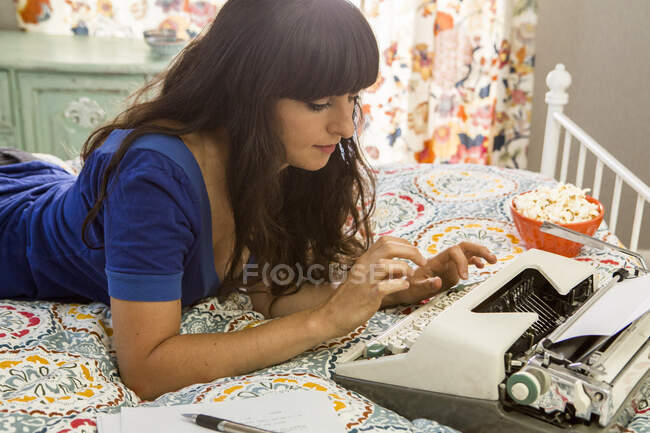 Jeune femme couchée sur le lit, à l'aide d'une machine à écrire — Photo de stock