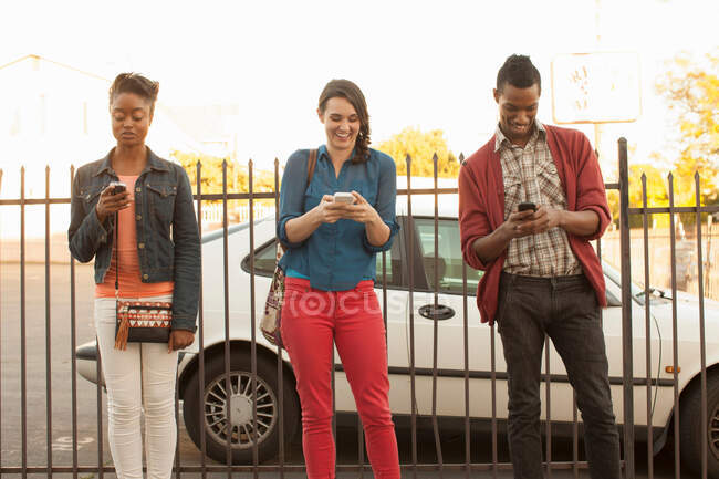 Tres amigos usando teléfonos celulares por valla metálica - foto de stock