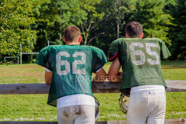 Vista posteriore di due giocatori di football americano maschi adolescenti appoggiati sulla recinzione del campo da gioco — Foto stock