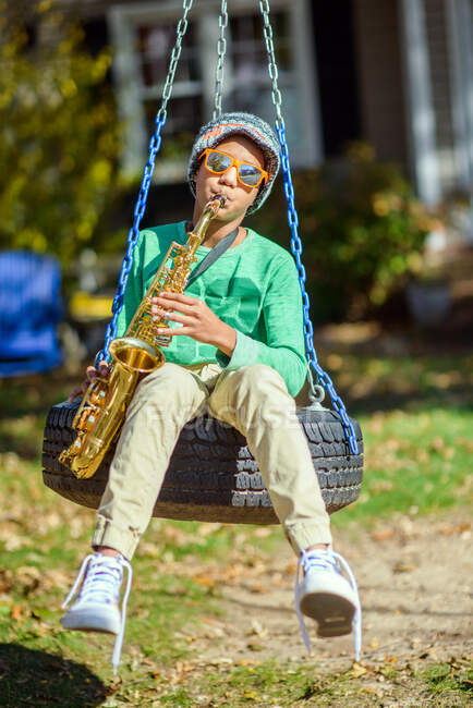 Adolescent garçon, assis sur la balançoire des pneus, jouer au saxophone — Photo de stock