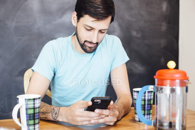 Молодой человек сидит за столом, пьет кофе, смотрит на смартфон — стоковое фото