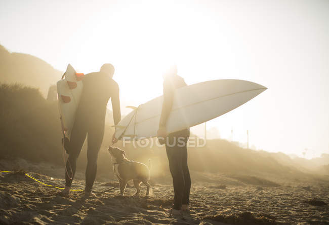 Surfers with dog in sunlight holding surfboards on beach, Malibu, California, USA — Stock Photo