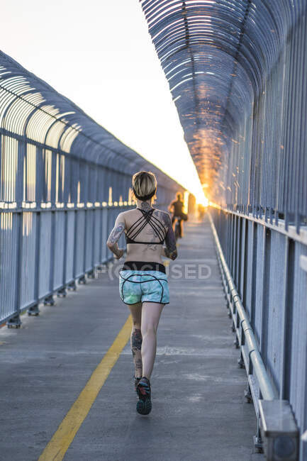 Jeune femme tatouée courir sur le pont avec coucher de soleil derrière — Photo de stock