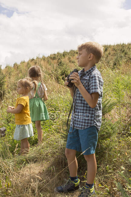 Tres niños pequeños explorando, al aire libre, niño usando prismáticos - foto de stock