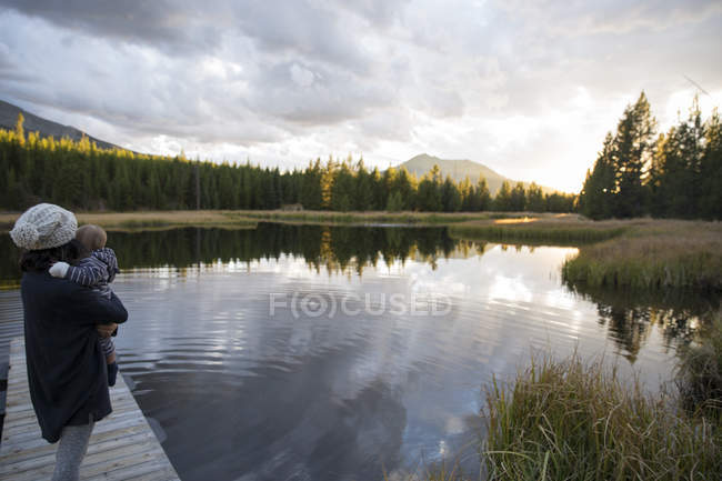 Mãe de pé no cais de madeira ao lado do lago, segurando o filho jovem — Fotografia de Stock