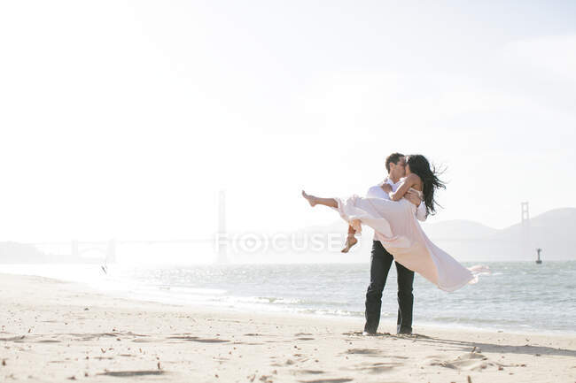 Romantic man carrying girlfriend in arms on beach, San Francisco Bay, USA — Stock Photo