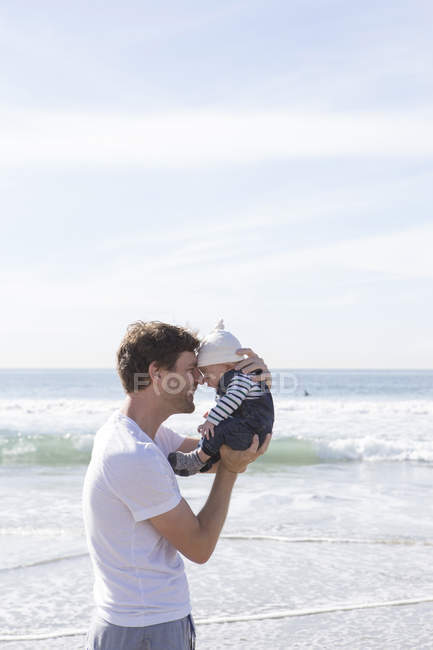 Father holding baby boy, face to face, at beach — Stock Photo
