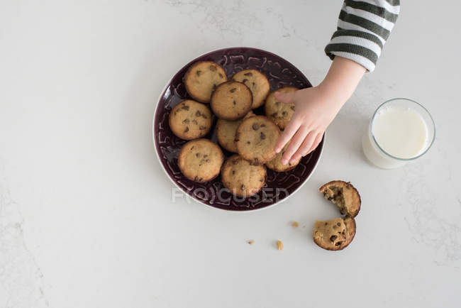 Imagen recortada de niño tomando galletas recién horneadas del plato, vista aérea, primer plano - foto de stock