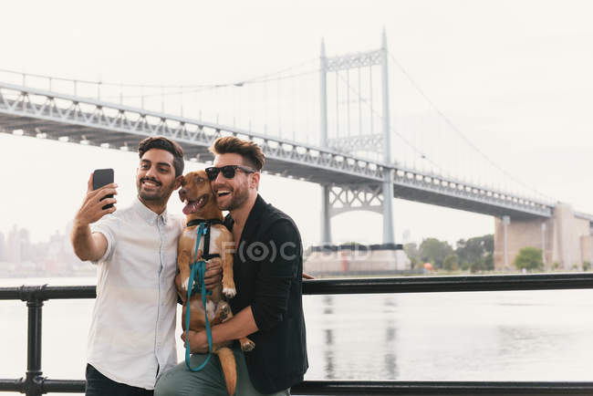 Joven pareja masculina a orillas del río tomando selfie de smartphone con perro, Astoria, Nueva York, Estados Unidos - foto de stock