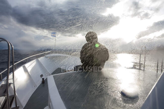 Человек в гидроизоляции, сидящий перед лодкой, плывущей под дождем, Ушуайя, Огненная Земля, Аргентина — стоковое фото