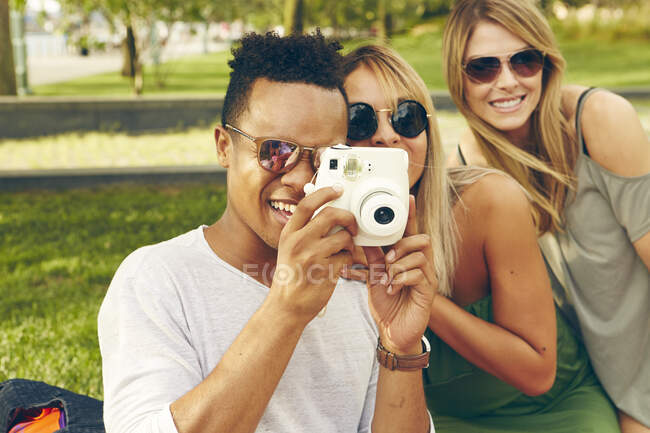 Молодой человек и друзья фотографируются с помощью мгновенной камеры в парке — стоковое фото