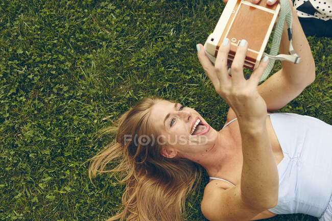 Молода жінка лежить на траві бере селфі з ретро камерою — стокове фото