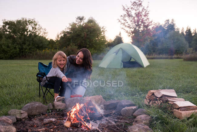 Madre e figlia sedute accanto al falò, a cucinare salsicce sul fuoco — Foto stock