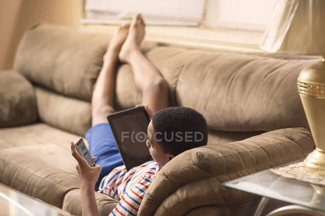 Jovem menino reclinado no sofá usando tablet eletrônico e smartphon — Fotografia de Stock