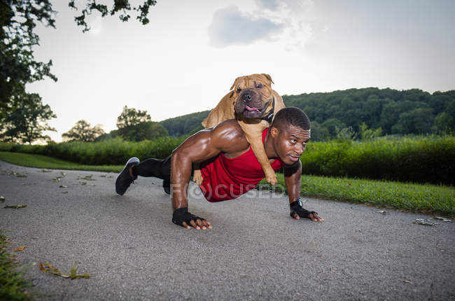 Jovem fazendo flexões na estrada rural, dando ao cão um piggyback — Fotografia de Stock