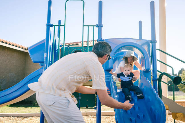 Família no parque infantil, o jovem filho deslizando para baixo slide — Fotografia de Stock