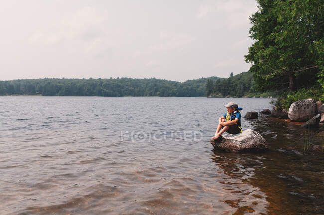 Niño sentado en la roca en el borde de un lago - foto de stock
