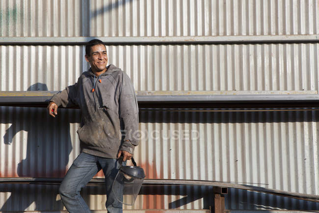 Bauarbeiter lehnt an Wellblechgebäude, schaut weg und lächelt — Stockfoto