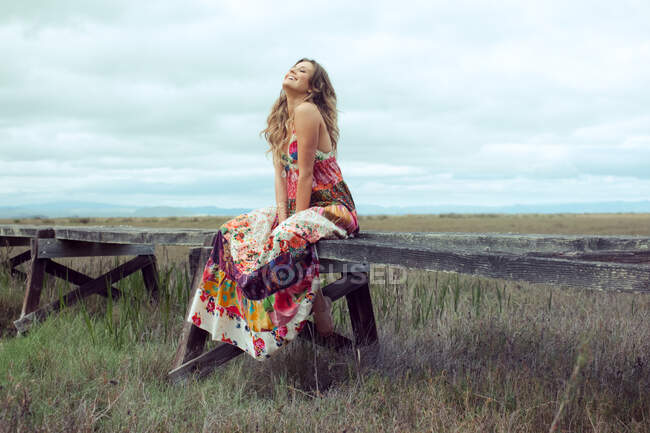 Молодая женщина в макси-платье сидит на возвышенной деревянной дорожке в пейзаже — стоковое фото