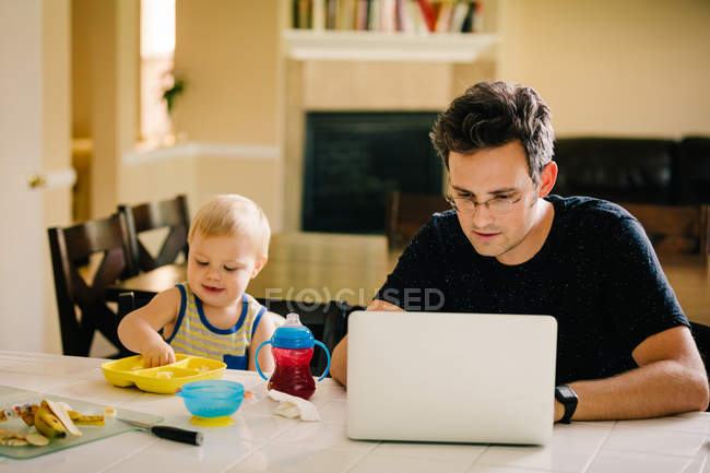 Father and young son sitting at table, son eating, father using laptop — Stock Photo