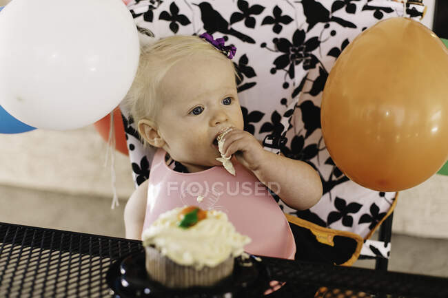 Baby girl sitting in high chair, eating birthday cake — Stock Photo