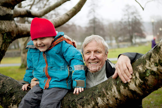 Abuelo y niño sentado en la rama del árbol, sonriendo - foto de stock