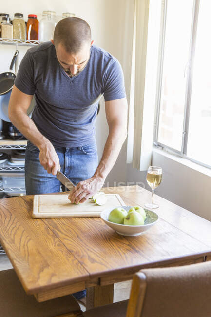 Середній дорослий чоловік нарізає яблуко за кухонним столом — стокове фото