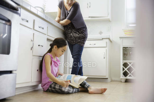 Menina sentada no chão da cozinha e livro de leitura — Fotografia de Stock
