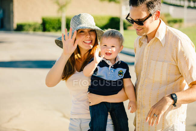Familia joven de pie al aire libre, madre e hijo pequeño saludando - foto de stock