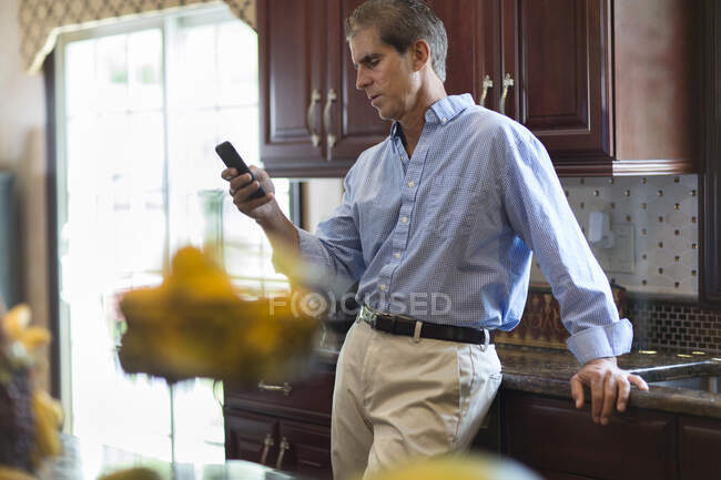 Мужчина средних лет на кухне смотрит на смартфон — стоковое фото