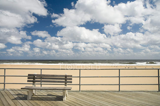 Bench on boardwalk with sandy beach and cloudy sky — Stock Photo