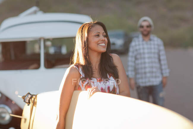 Mujer joven sosteniendo tabla de surf en el viaje por carretera, sonriendo - foto de stock