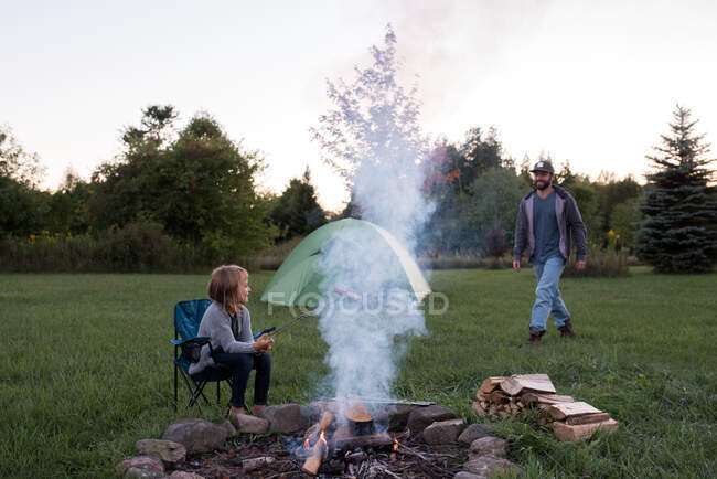 Young girl cooking sausage over campfire, father walking towards her — Stock Photo