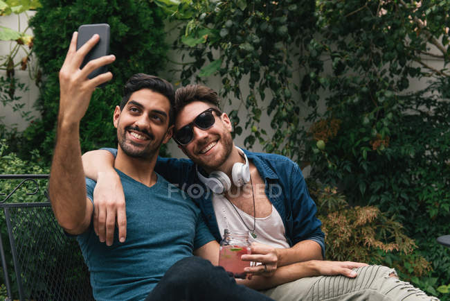Jovem casal masculino sentado no jardim e levando selfie smartphone — Fotografia de Stock
