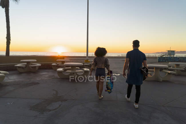 Couple walking near beach, holding skateboards, rear view — Stock Photo