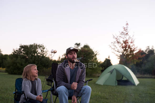 Padre e hija en sillas de camping, comiendo malvaviscos tostados - foto de stock