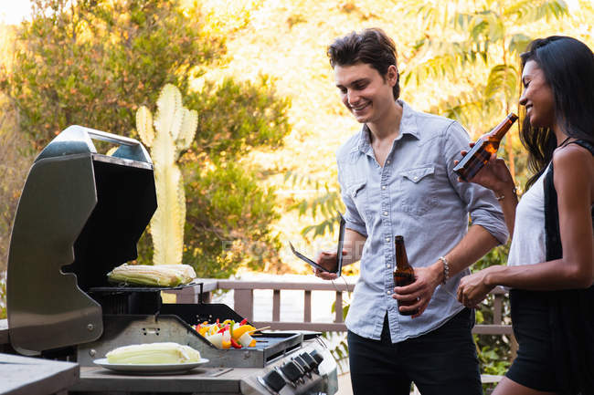 Jovem casal ao ar livre, segurando garrafas de cerveja, cozinhar alimentos em churrasco — Fotografia de Stock