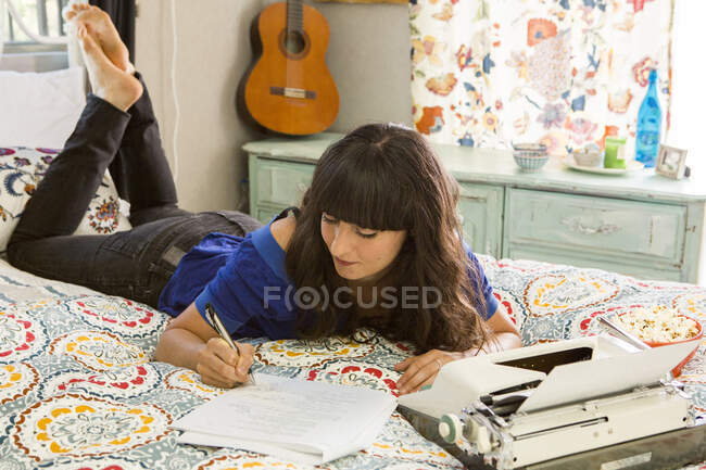 Mujer joven acostada en la cama, usando máquina de escribir, escribiendo notas - foto de stock