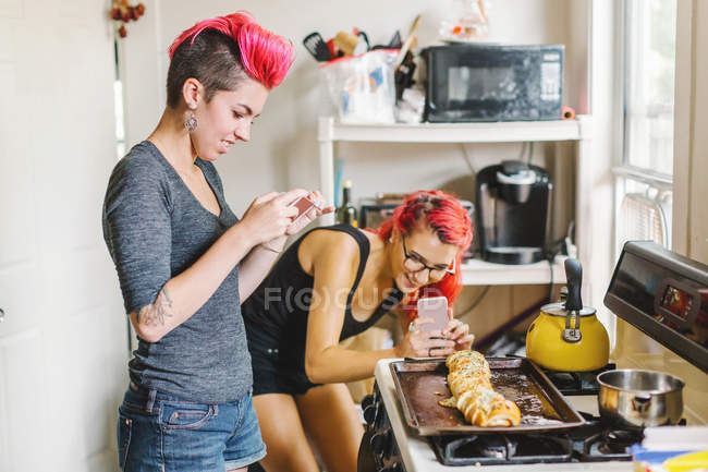Две молодые женщины с розовыми волосами фотографируют на смартфоне фаршированный багет на кухне — стоковое фото