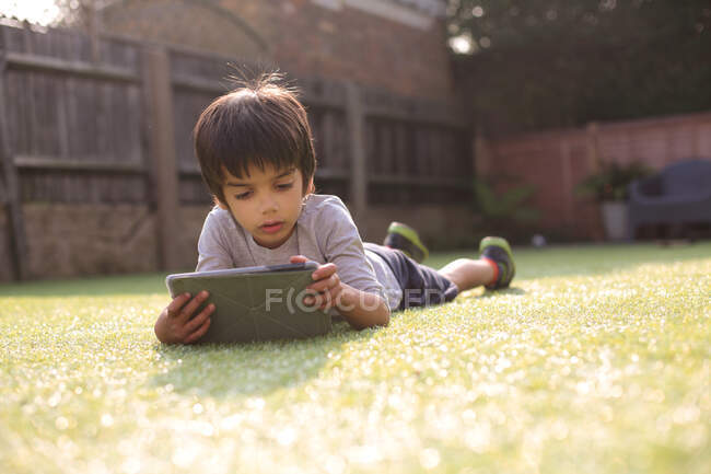 Мальчик в саду лежит на траве и смотрит вниз с помощью цифрового планшета — стоковое фото