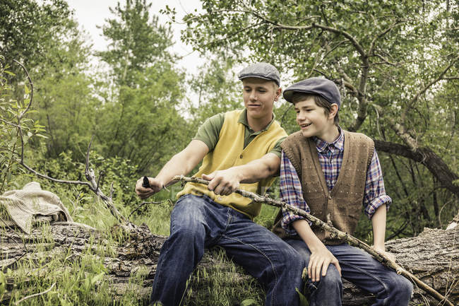 Man and boy wearing flat caps sitting on fallen tree whittling twig with penknife — Stock Photo