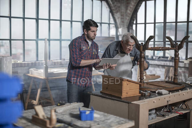 Cape Town, South Africa, young co worker reading off his pad to elderly craftsman while working on wooden chair — Stock Photo
