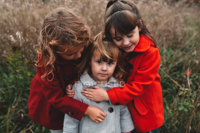 Retrato de dos niñas abrazando a su hermana pequeña en el campo - foto de stock
