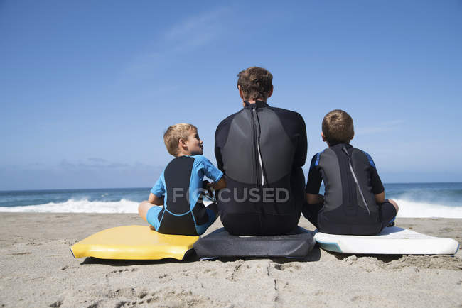 Rear view of man and two sons sitting on bodyboards, Laguna Beach, California, USA — Stock Photo