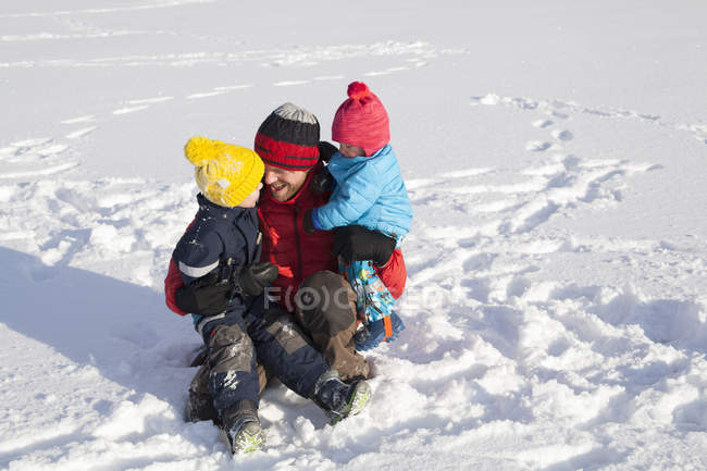 Padre sentado en la nieve, abrazando a dos hijos - foto de stock